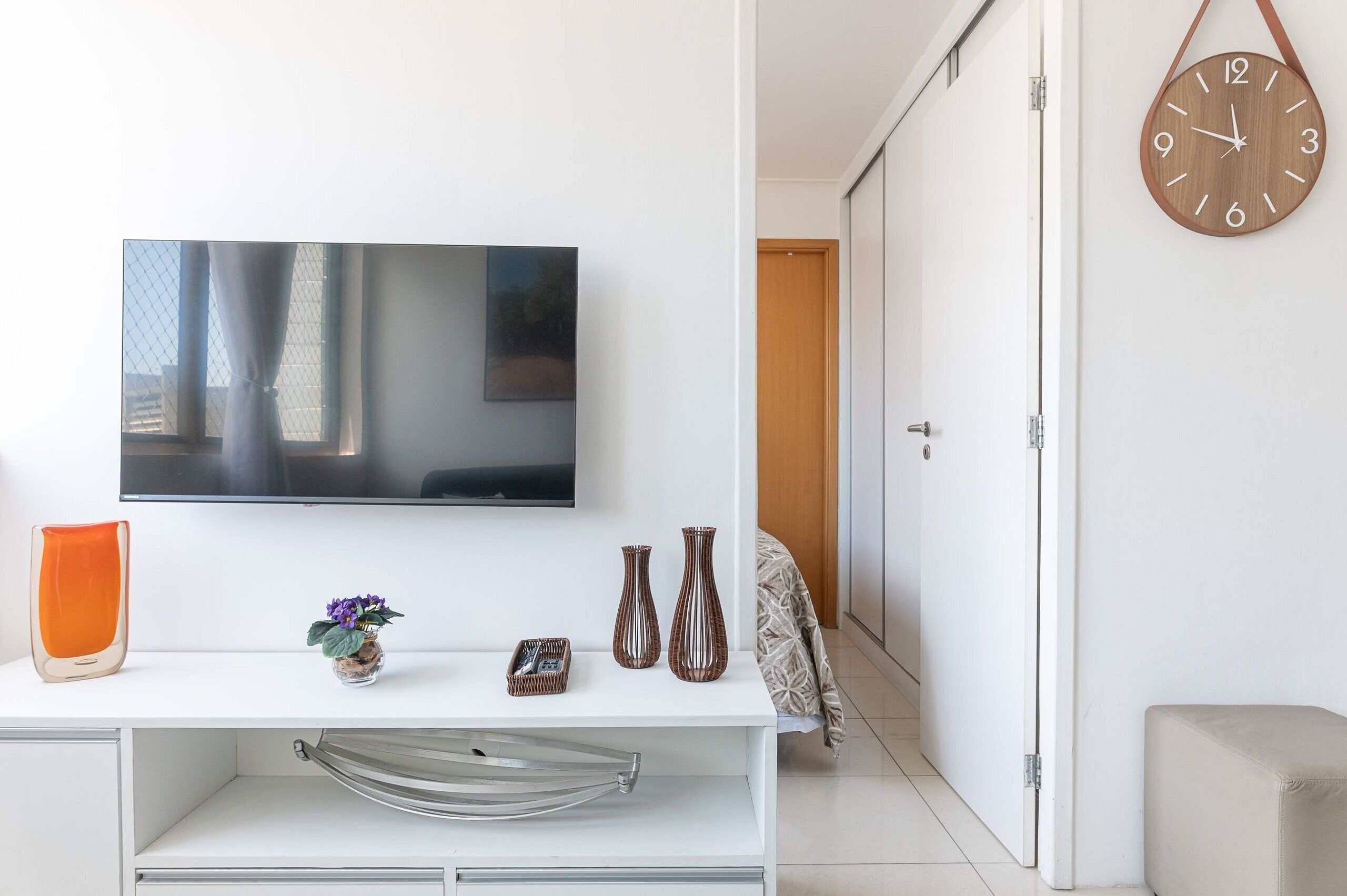 A modern living room with a flat-screen TV mounted on the wall, a white console table with decorative items, and a wooden clock on the wall.