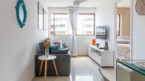 A modern living room with a grey sofa, a small side table with a vase, a white ceiling fan, and a large window with curtains. - 109 Flat completo e acolhedor nas Graças (Recife)