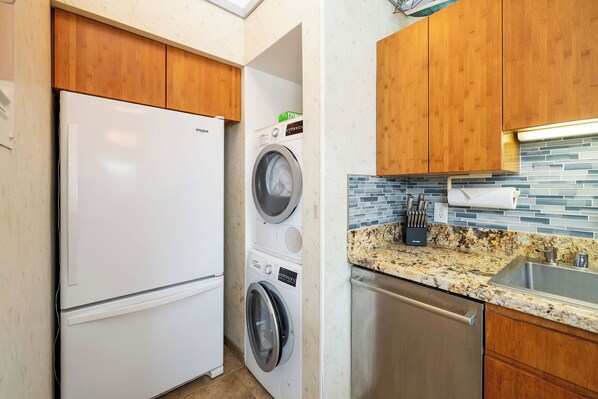 A compact kitchen with a refrigerator, washing machine, dishwasher, and a granite countertop with a sink. - Ocean view with Loft (Kailua-Kona)