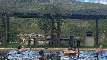 Una piscina con gente disfrutando el agua, rodeada de un paisaje natural y una montaña al fondo.