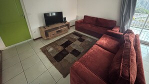 A living room with a TV on a wooden stand, a red sofa, a patterned rug, and a green door. - O refuge d'Hell-Bourg (Salazie)