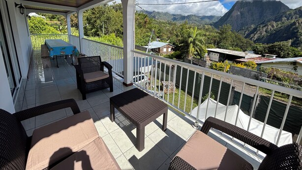 A covered outdoor area with wicker furniture, a table, and chairs. It overlooks a mountainous landscape with greenery and buildings. - O refuge d'Hell-Bourg (Salazie)