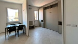 A room with a dining table and chairs, a large wardrobe, a radiator, and a window with a view of buildings. - CN014 - Cesenatico, new three-room apartment on the canal port (Cesenatico)