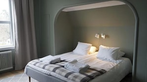 Una cama bien hecha con una colcha y almohadas de cuadros, un radiador y una ventana con cortinas.