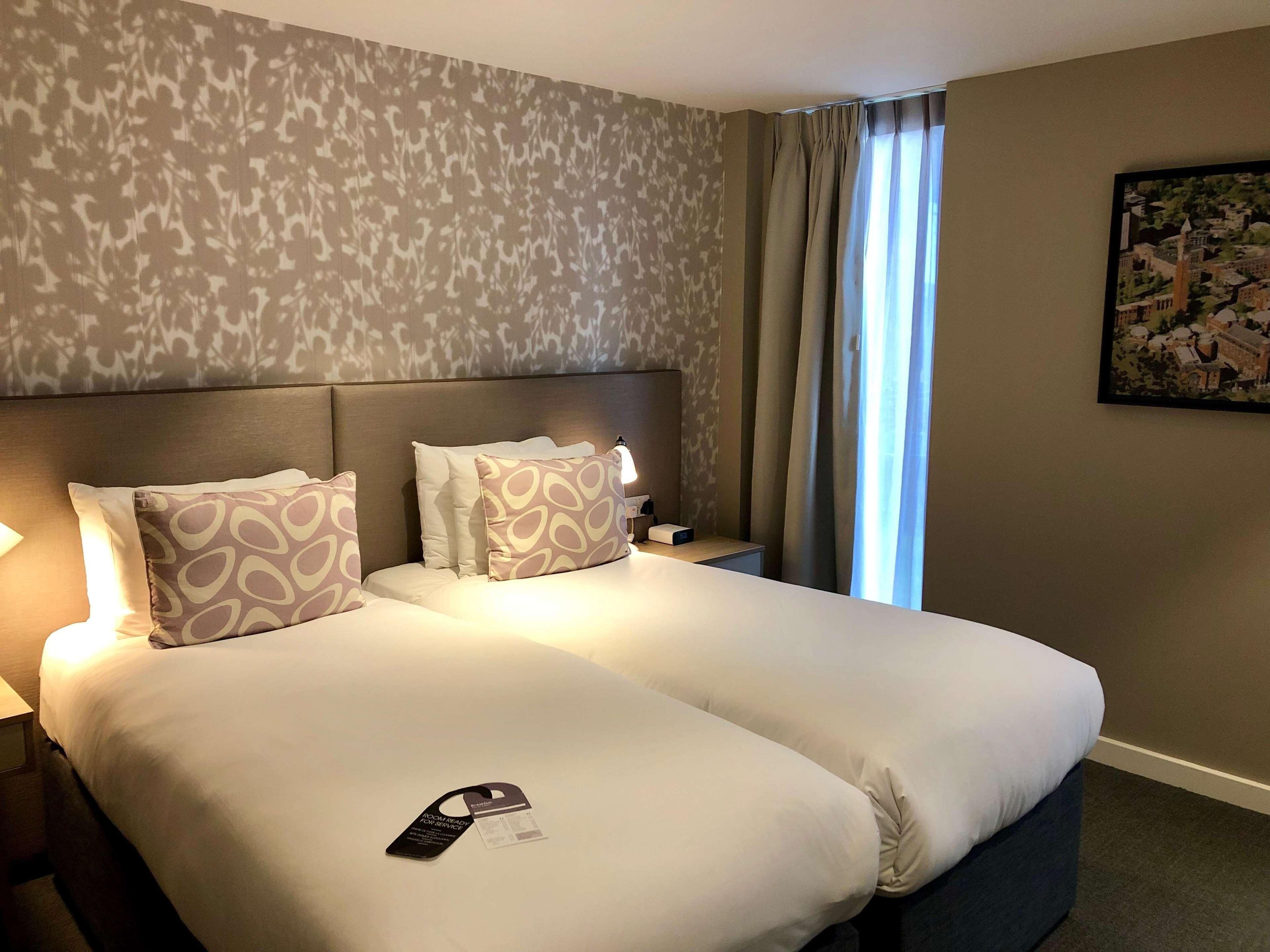 Habitación de hotel con cama doble, almohadas estampadas, mesita de noche con lámpara, un cuadro en la pared y ventana con cortinas.