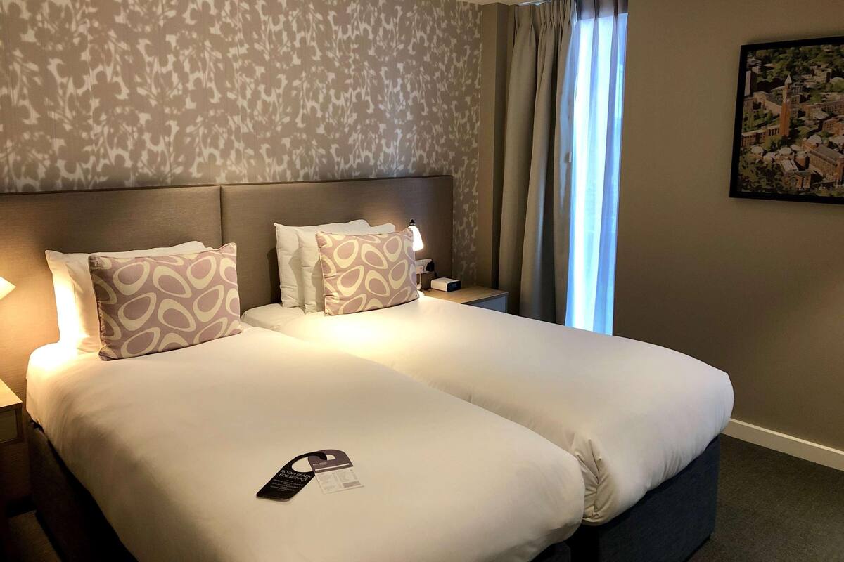 Habitación de hotel con cama doble, almohadas estampadas, mesita de noche con lámpara, un cuadro en la pared y ventana con cortinas.