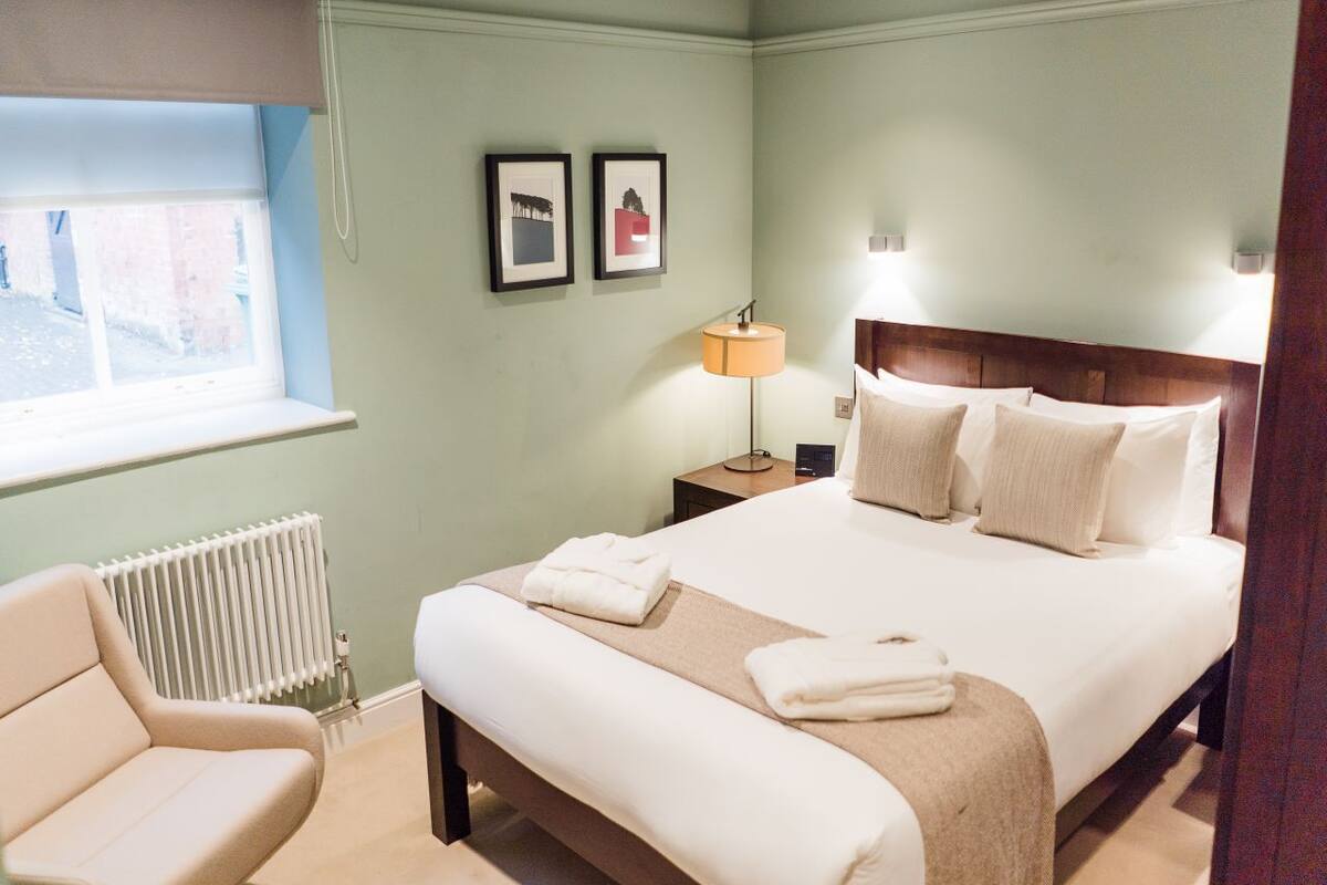 Un dormitorio ordenado con cama, lámpara de noche y una silla.
