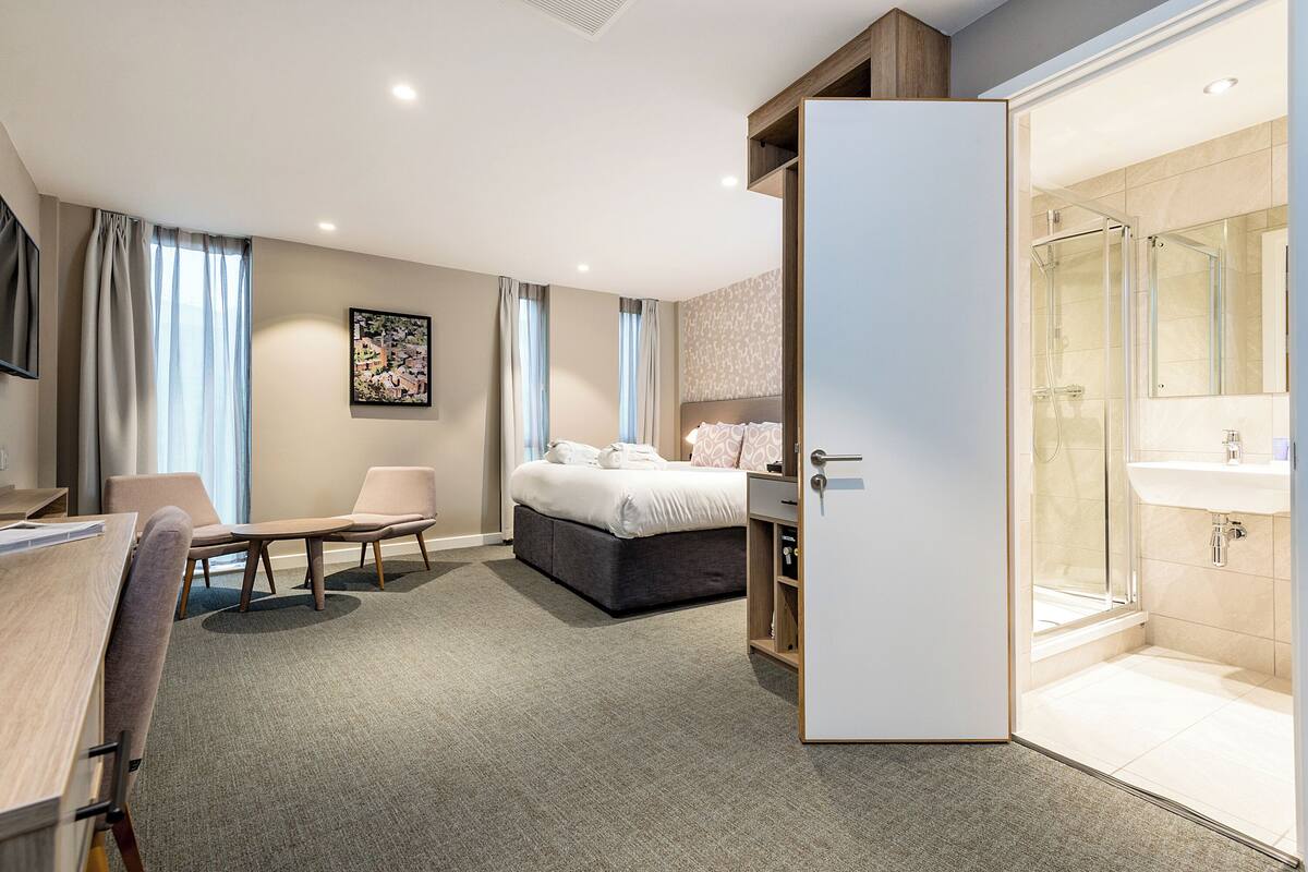 Habitación de hotel con cama, escritorio, silla, armario y baño con ducha y lavamanos.