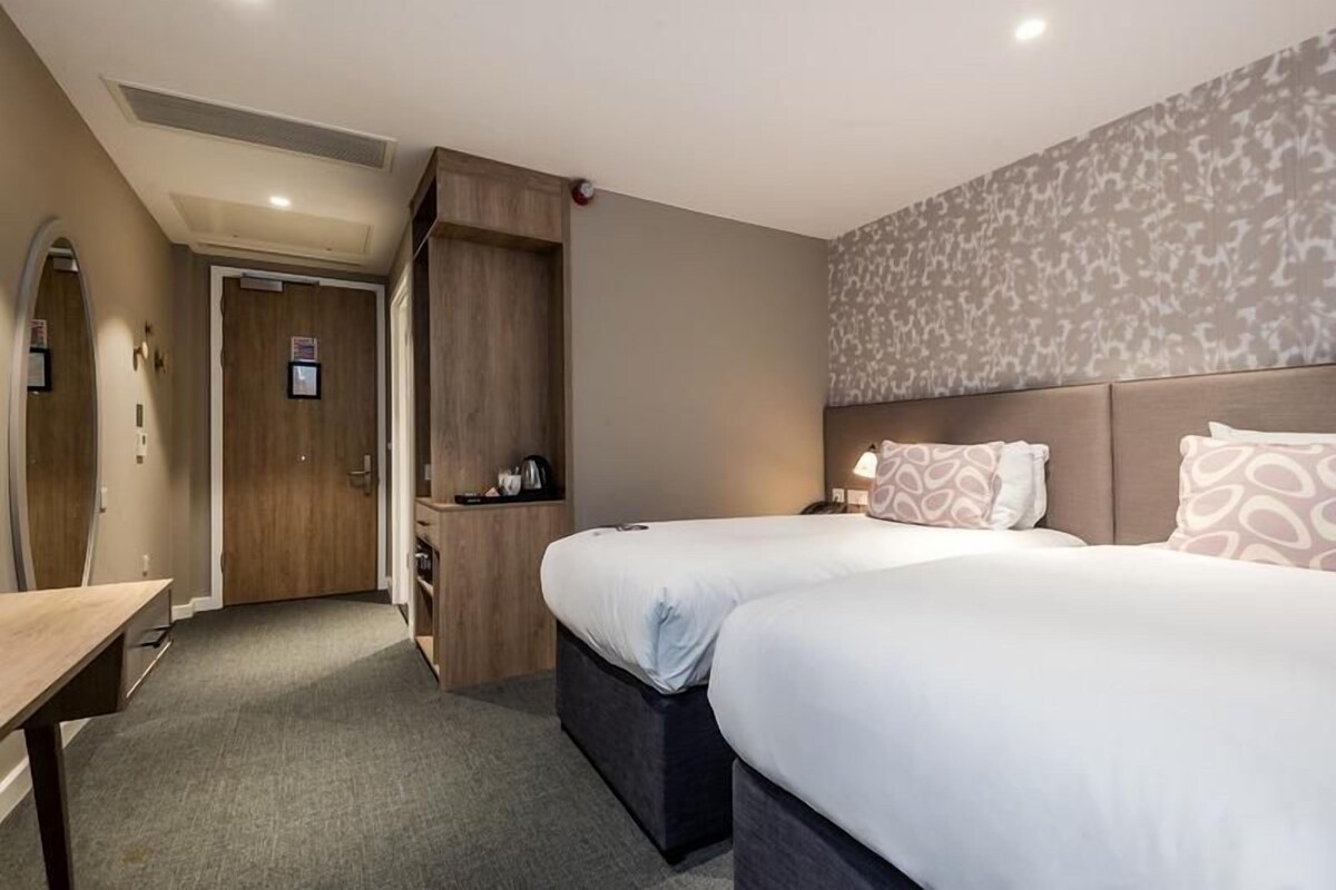 A hotel room with two beds, a desk, a wardrobe, and a shower area.