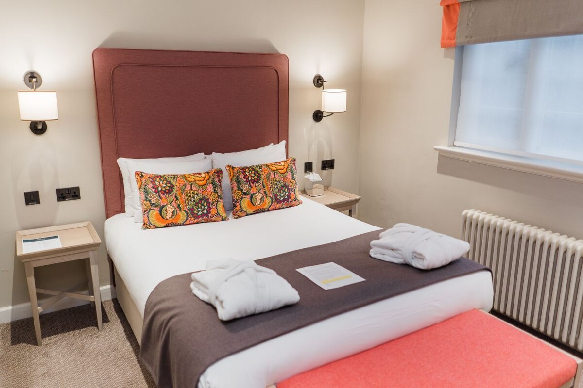 A neatly made bed with pillows, a bedside table, a nightstand, a window with a view, and a radiator.