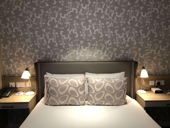 A hotel room with a bed, two bedside lamps, a nightstand with a phone, a digital clock, and a patterned wall. - Edgbaston Park Hotel & Conference Centre (Birmingham)