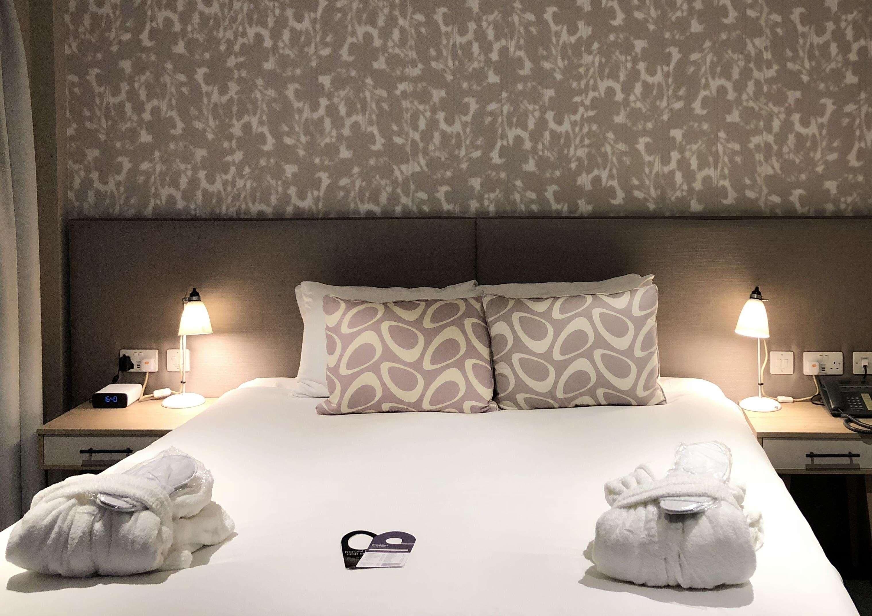 Una habitación de hotel con una cama, dos lámparas de noche, una mesita de noche con un teléfono y una lámpara, y dos toallas sobre la cama.
