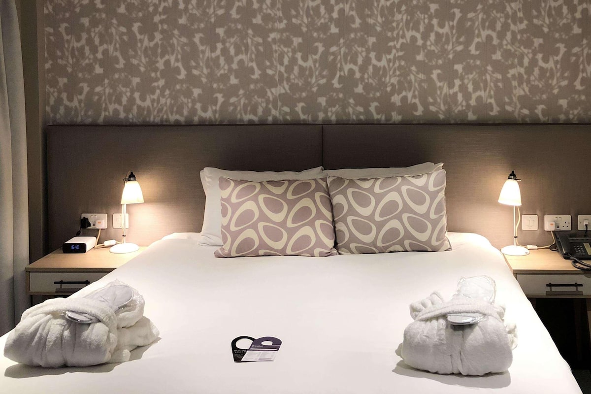 Una habitación de hotel con una cama, dos lámparas de noche, una mesita de noche con un teléfono y una lámpara, y dos toallas sobre la cama.