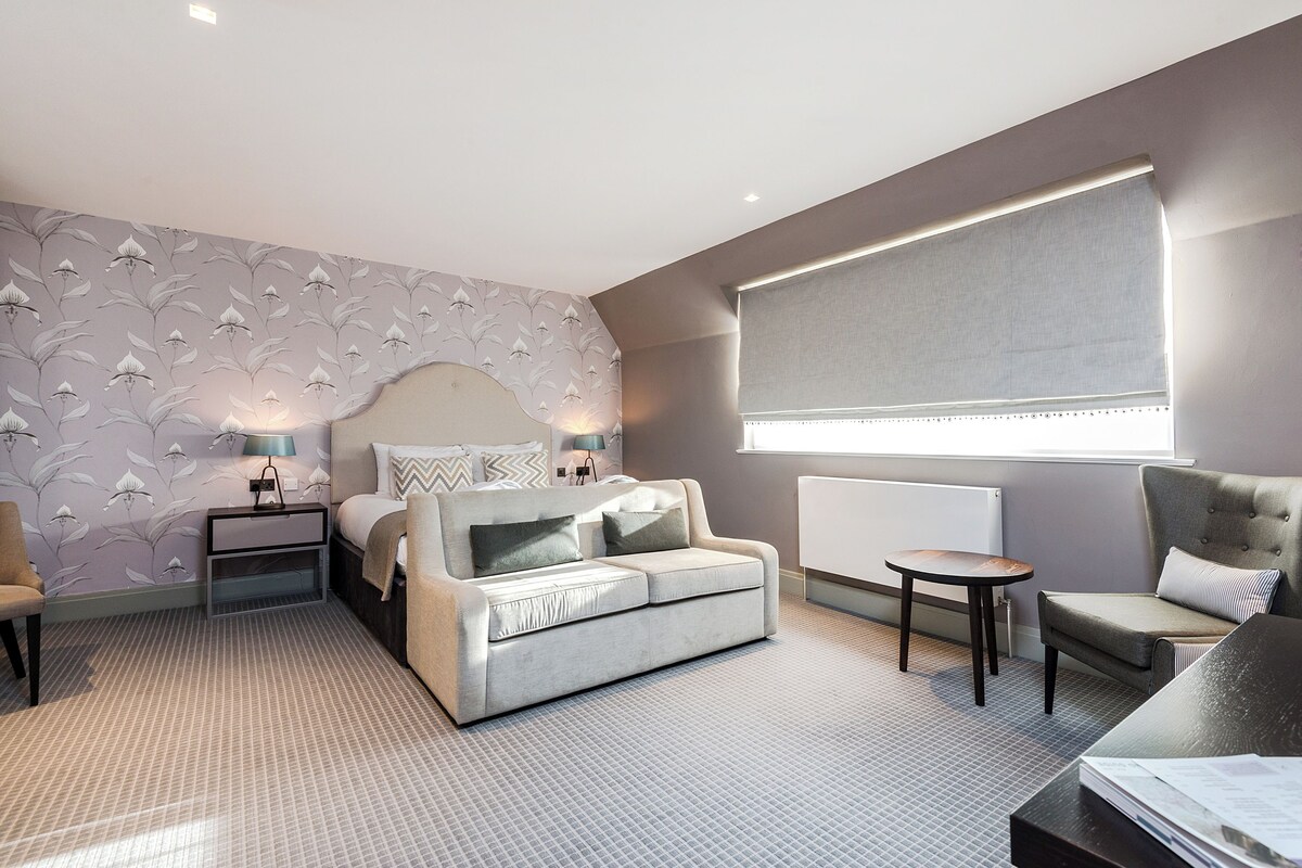 A modern hotel room with a sofa, a chair, a small table, a nightstand, and a lamp.