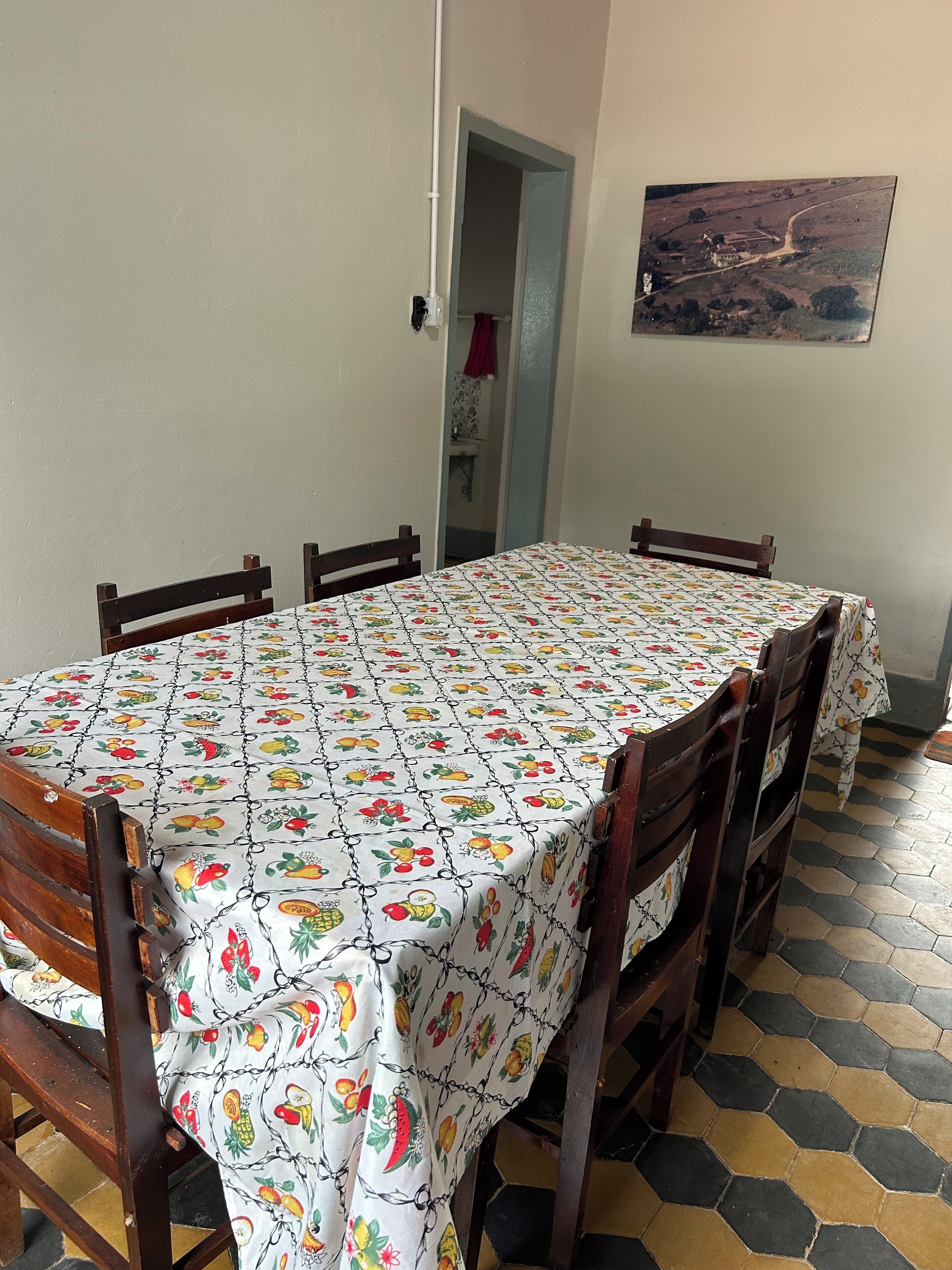 A dining area with a table covered by a fruit-patterned tablecloth, surrounded by wooden chairs.