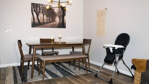 A dining area with a wooden table, chairs, a bench, a chandelier, a macrame wall hanging, and a baby rocker.