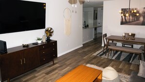 A modern living room with a flat-screen TV, wooden furniture, a ceiling fan, and a dining area with a table and chairs.