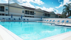 A swimming pool with lounge chairs and umbrellas, surrounded by a white fence, with a building in the background.