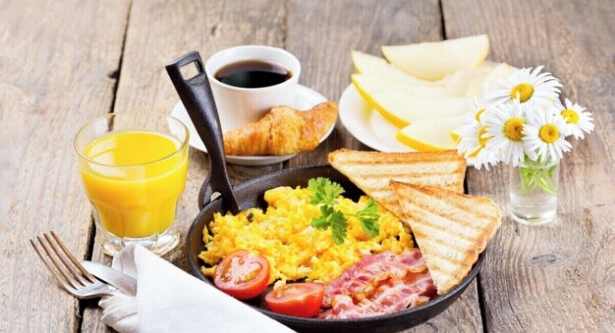 Une assiette de petit-déjeuner comprenant des œufs, du bacon, des tomates, des toasts et un verre de jus d’orange.