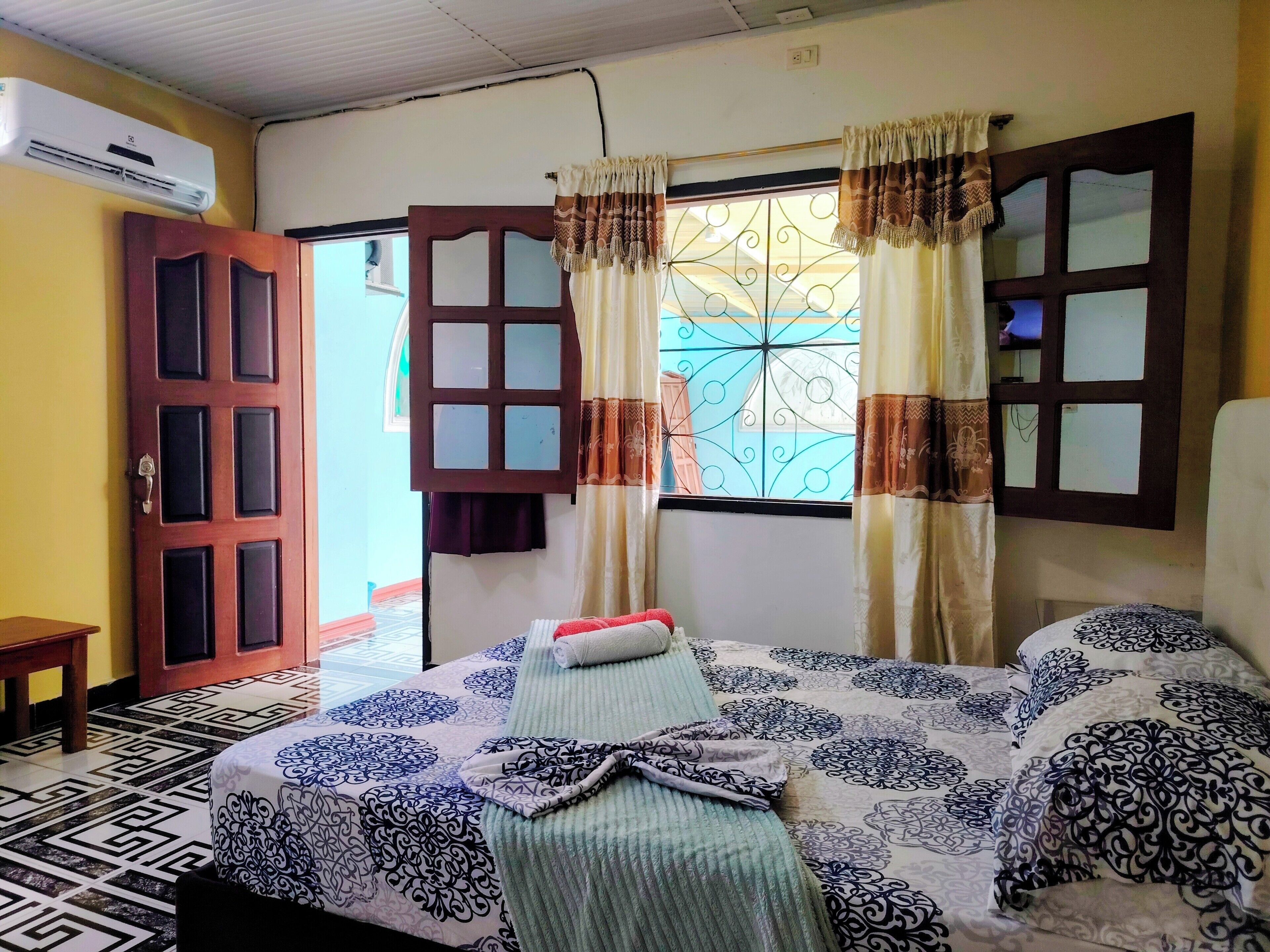 Una habitació amb un llit, una porta de fusta, un aparell d'aire condicionat i una finestra amb cortines decoratives.