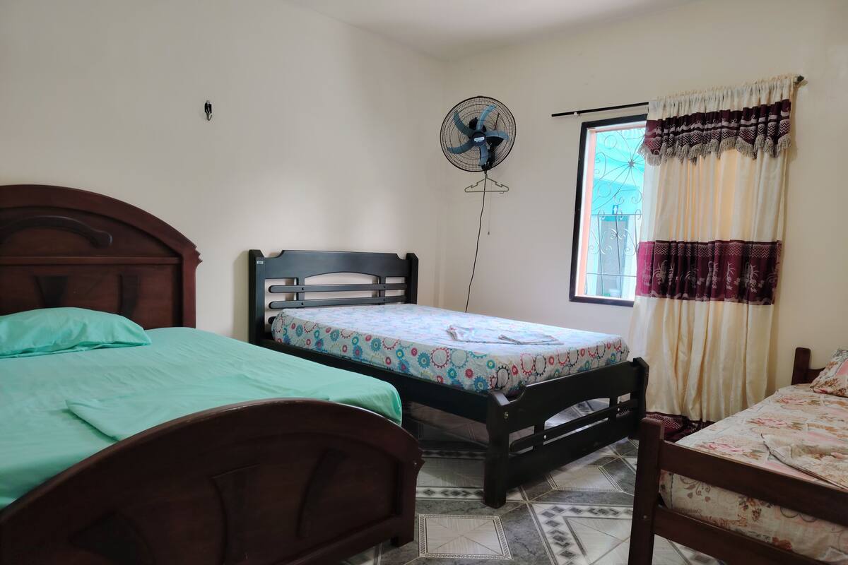 A bedroom with two wooden beds, a standing fan, a window with curtains, and a tiled floor.