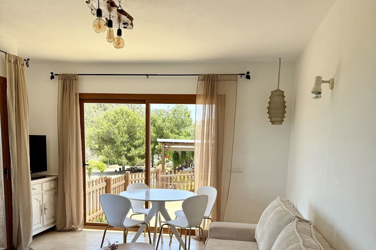 Un salon comprenant un canapé, une table basse et une table à manger avec des chaises. On y trouve également un téléviseur posé sur un meuble, et de grandes fenêtres offrent une vue sur l’extérieur.