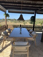 Un espacio cubierto al aire libre con mesa y sillas, una planta colgante y vistas del paisaje.
