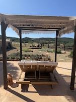 Un banco de madera con cojines bajo un techo de paja, con un paisaje seco y montañoso de fondo.