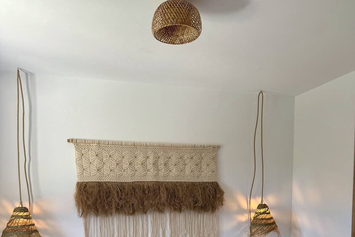 Une chambre spacieuse avec un grand lit, une décoration murale en macramé et deux lampes suspendues.