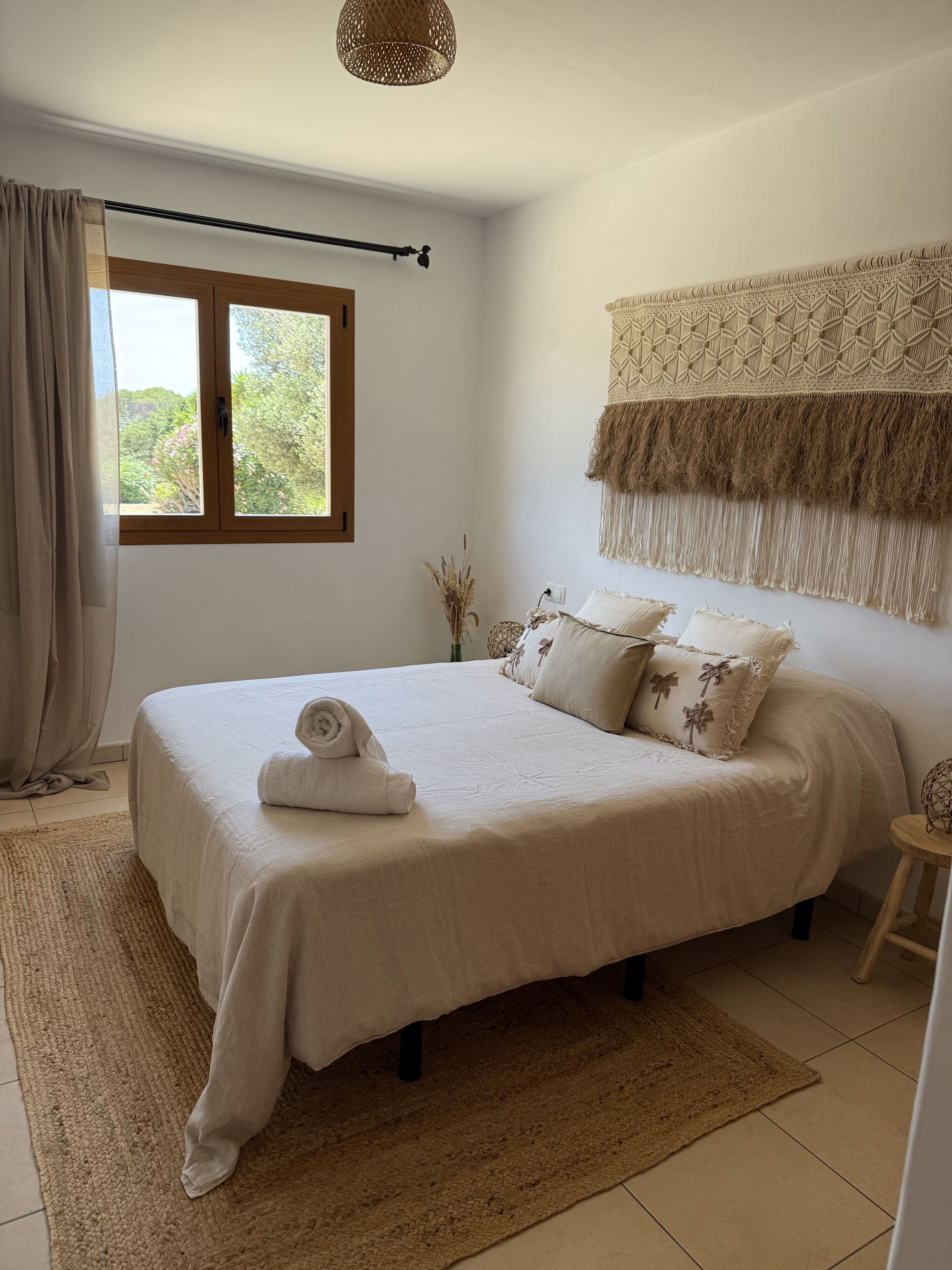 Ein Schlafzimmer mit einem großen Bett, einem Fenster mit Vorhängen, einem Wandbehang aus Makramee und einem kleinen Hocker.