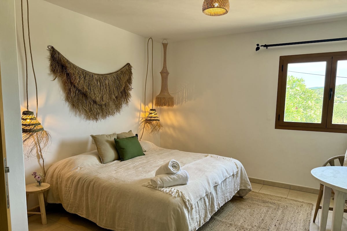 Une chambre à coucher avec un lit, des tables de chevet, une lampe suspendue, une fenêtre et une vue sur la verdure à l’extérieur.