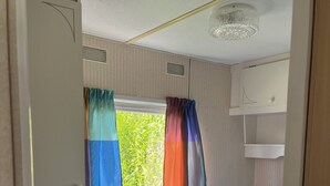 A compact bedroom with a single bed, a window with colorful curtains, a ceiling light fixture, and a small white cabinet. - ZP 360 - Camping de Zandput (Vrouwenpolder)