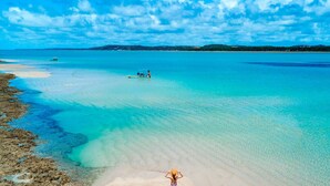 Une plage aux eaux turquoise et limpides, un bateau à l’horizon et une personne debout sur le sable.