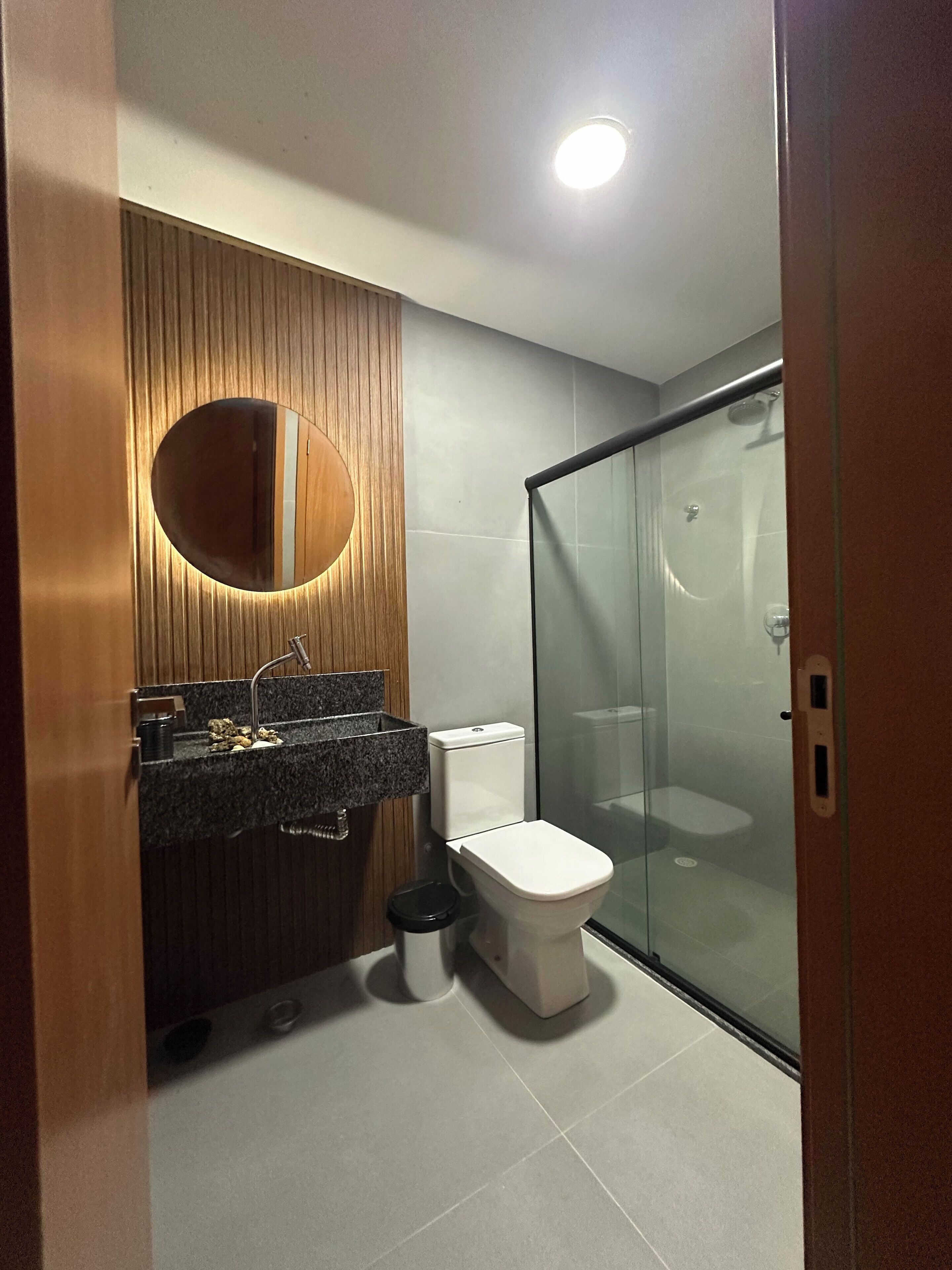 Un baño moderno con tocador de madera, un espejo redondo, un inodoro y una cabina de ducha con paredes de vidrio.