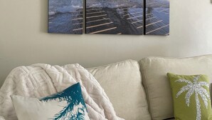 Ein Zimmer mit einem Bett, das mit einer weißen Decke bezogen ist, einem beigefarbenen Sofa und einem Wandbild von einem Pier über dem Meer.