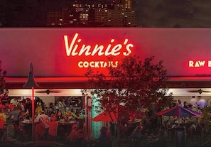 A bar area with tables and chairs, a large illuminated sign reading 'Vinnie's Cocktails,' and a city skyline in the background. - Tropical Paradise by the Sea - Huge Pool,BBQ,Beach,FIFA World Cup & 4th of July! (Lauderdale-by-the-Sea)