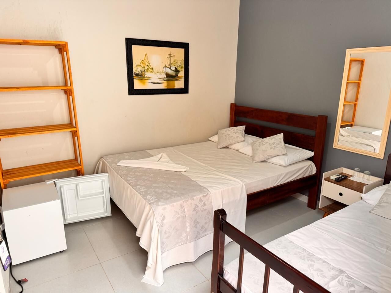 Un dormitorio con una cama, cabecera de madera, mesita de noche, espejo, un cuadro en la pared y un pequeño armario blanco.