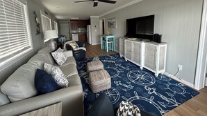Ein Wohnzimmer mit einer Couch, einem Hocker und einem Fernseher auf einem Schrank. Es gibt einen Deckenventilator, eine Küche im Hintergrund und einen Essbereich mit Tisch und Stühlen.