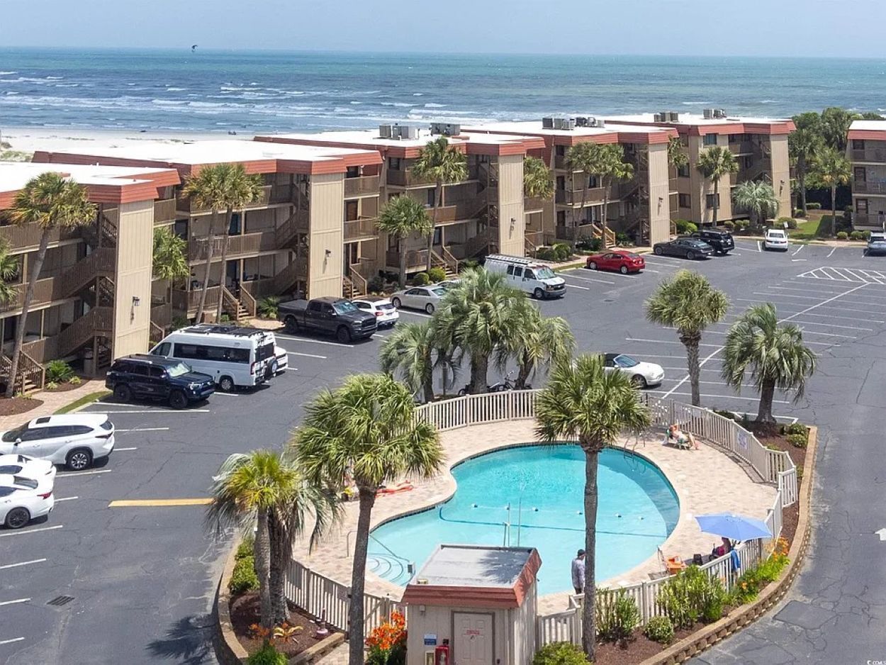 A hotel with a pool surrounded by palm trees, a parking lot with several cars, and a view of the ocean.