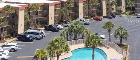 A hotel with a pool surrounded by palm trees, a parking lot with several cars, and a view of the ocean.