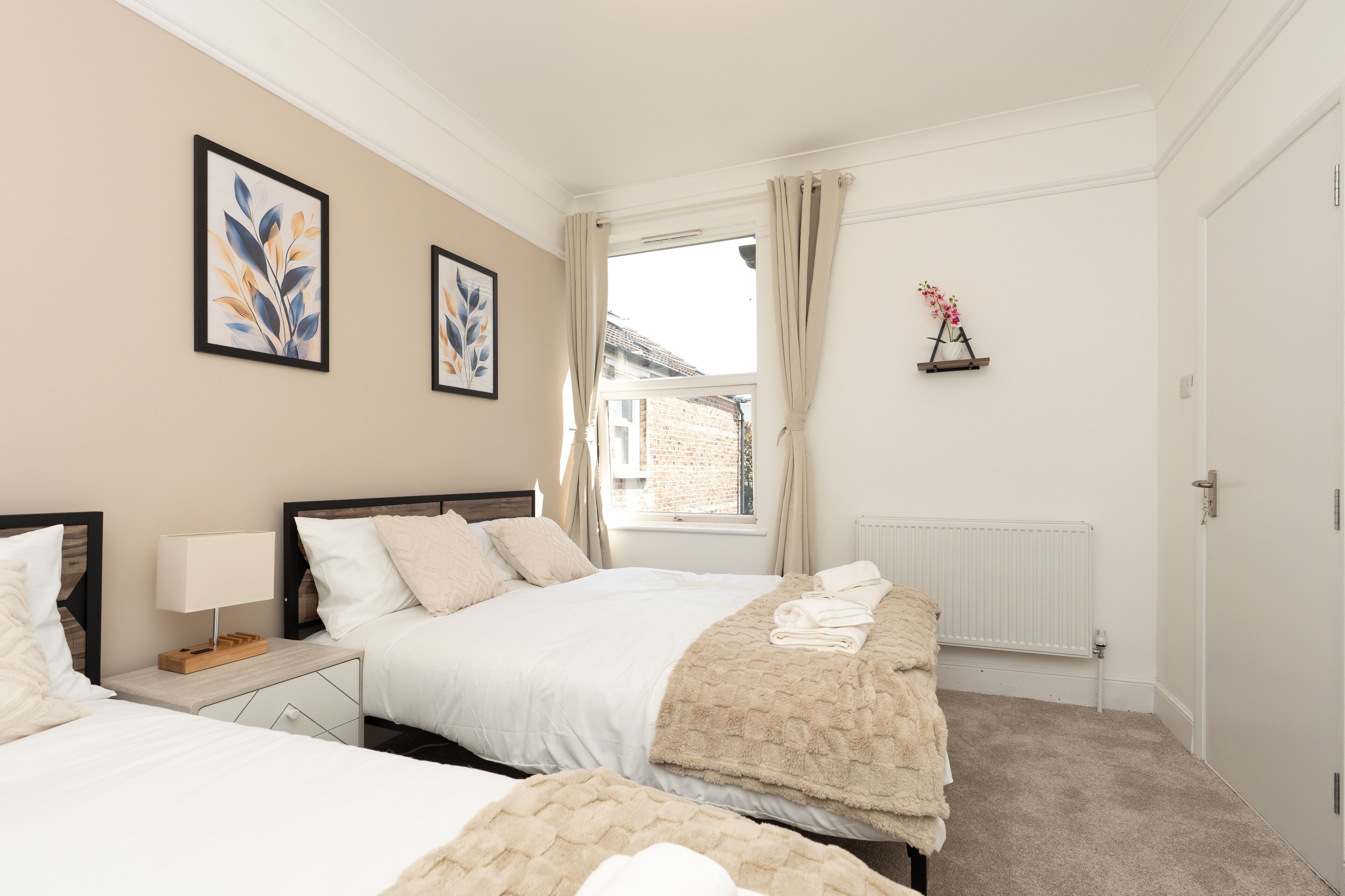 Une chambre avec deux lits, une fenêtre avec des rideaux, un radiateur et des œuvres d’art encadrées accrochées au mur.