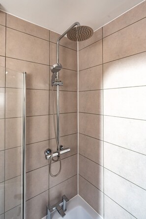 Une douche moderne dotée d’un pommeau de douche à main et d’une grande pomme de douche au plafond, le tout installé sur un mur recouvert de grands carreaux carrés.