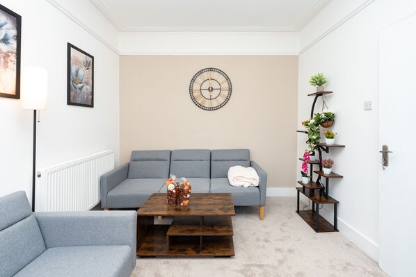 Un salon avec un canapé gris, une table basse, une horloge murale et une étagère pour plantes.