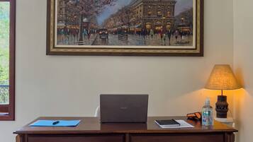 A room with a desk, two chairs, a laptop, a framed painting, a lamp, and a bottle of hand sanitizer.