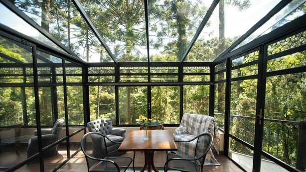 Een oranjerie met een houten tafel en stoelen, omringd door grote glasramen met uitzicht op hoge bomen.
