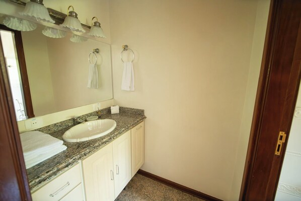 A bathroom with a granite countertop, white sink, and a mirror above the sink. There are two towels hanging on hooks and a toilet paper roll on the counter.