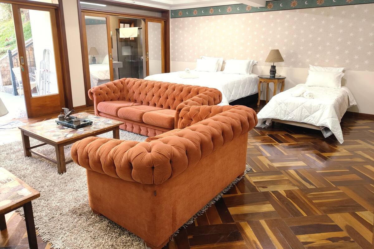 Hotellihuone, jossa on napitettulla kuviolla varustettu sohva, valkoisilla tekstiileillä varustettu sänky, pieni pöytä lampun kanssa ja kalanruotokuviollinen puulattia.