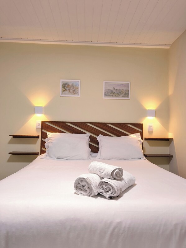 Una cama bien hecha con ropa de cama blanca y toallas enrolladas, dos mesitas de noche fijadas a la pared y dos cuadros enmarcados.