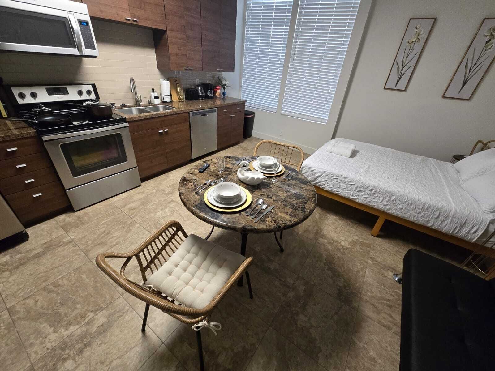 A compact kitchen with a stove, microwave, and sink. There's a dining table with chairs and a bed with a white comforter.