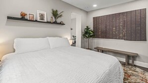 A bedroom with a large bed, a bench, a wall-mounted shelf with decorative items, and a large abstract artwork.